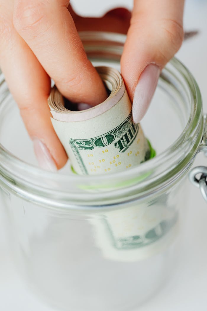 about-03 Hand placing rolled US dollars into a glass jar, representing savings and financial planning.